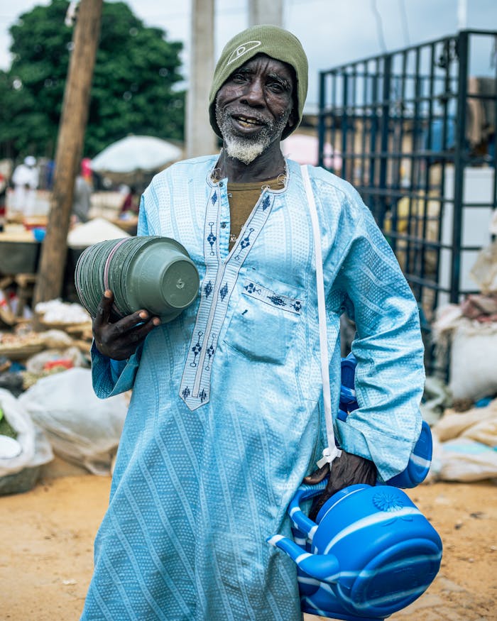 who-we-are An elderly man dressed in traditional attire holding household items at an outdoor market, smiling warmly.